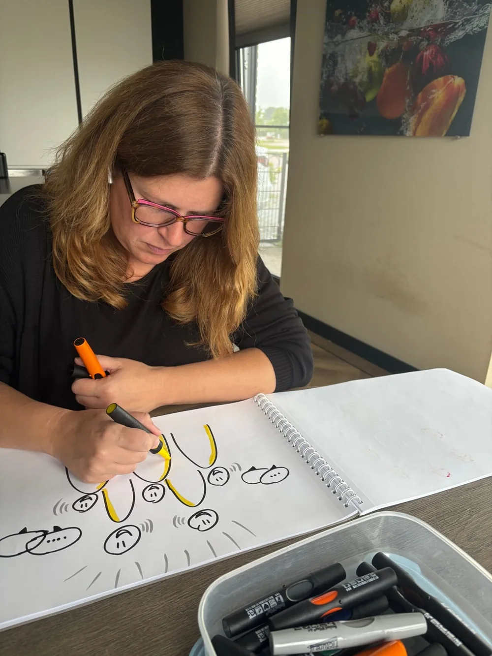 Foto van onze illustrator aan het werk: met viltstift tekent ze de gele poppetjes van Begrijpjekind.nl in een schetsboek, met een bak stiften naast zich.